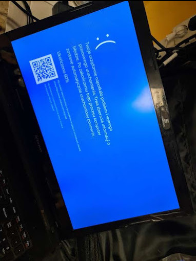 Ekran laptopa Lenovo wyświetlający niebieski ekran śmierci z kodem QR i komunikatem o błędzie, postęp ukończony w 60%, widoczne klawisze laptopa i fragment nieostrego tła.