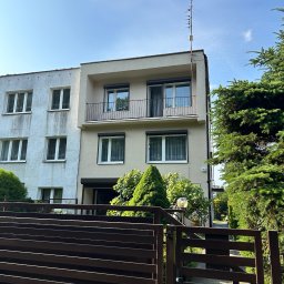 Firma Ogólnobudowlana Wiktor Marczak - Dwukondygnacyjny budynek z balkonem, z widoczną różnicą w kolorze elewacji na części budynku. Antena na dachu. Brązowe ogrodzenie na pierwszym planie.