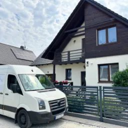 Dwukondygnacyjny dom z drewnianą elewacją i białymi ścianami, widziany zza szarego ogrodzenia. Na pierwszym planie biały bus z reklamą firmy elewacyjnej.