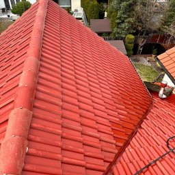 Firma Ogólnobudowlana Wiktor Marczak - Czerwona dachówka ceramiczna po czyszczeniu, widoczne ślady wilgoci. W tle zabudowa jednorodzinna i zieleń. Ujęcie z góry, lekki kąt.