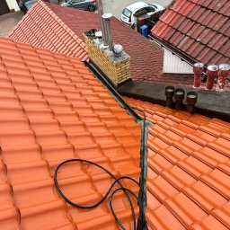 Firma Ogólnobudowlana Wiktor Marczak - Błyszcząca, pomarańczowa dachówka ceramiczna po myciu, widok z góry. W tle komin z cegły i fragmenty innych dachów. Widoczne przewody i lanca myjki ciśnieniowej.