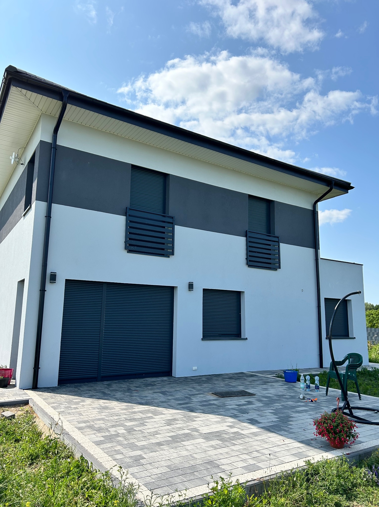 Dwukondygnacyjny dom z szarą elewacją i grafitowymi akcentami, widok z poziomu kostki brukowej. Na tarasie widoczne akcesoria, krzesło i kwiaty.