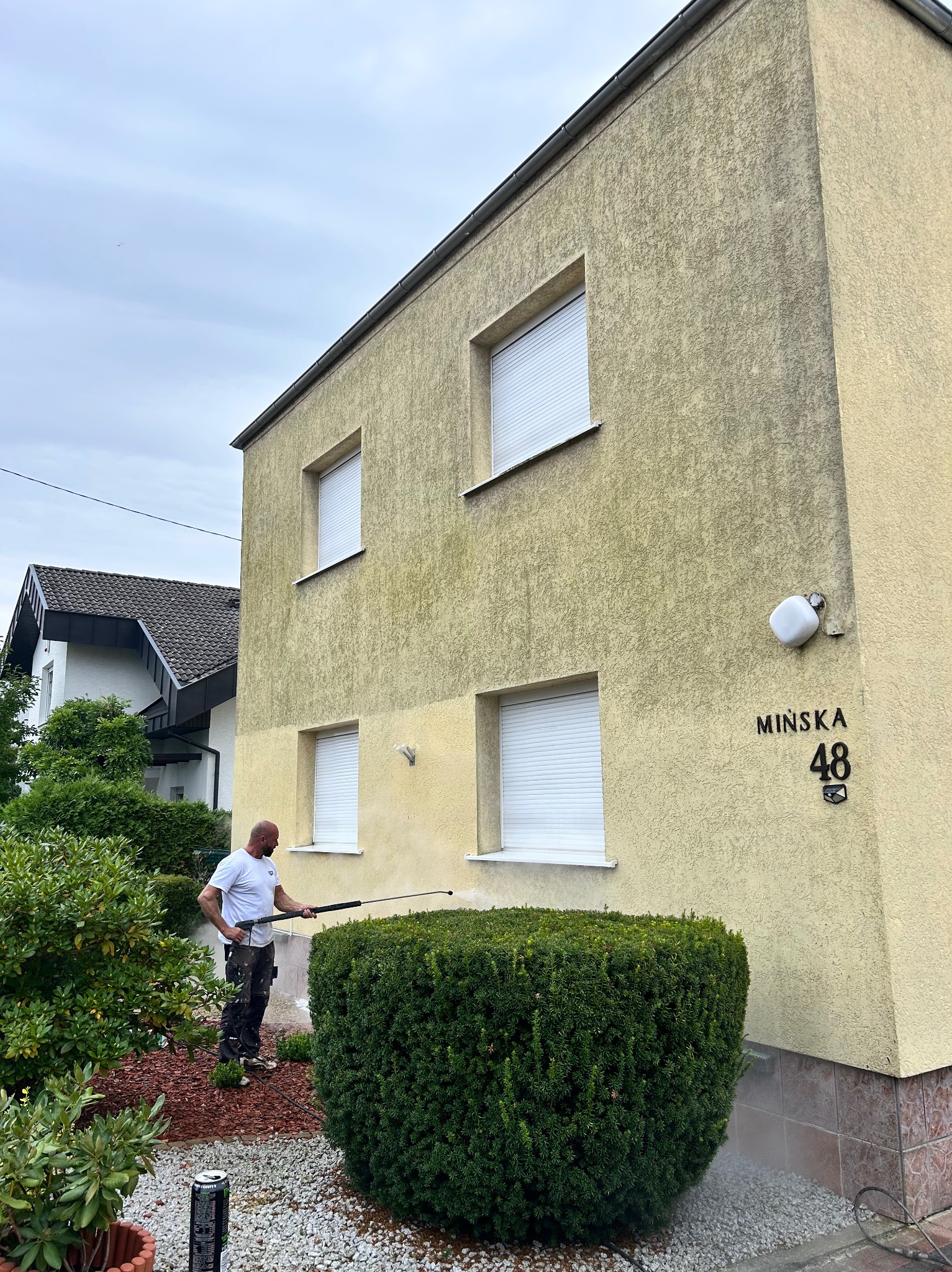 Czyszczenie elewacji domu o beżowej strukturze, widoczny fragment wyczyszczonej ściany, pracownik z lancą ciśnieniową i adres 'Mińska 48'.