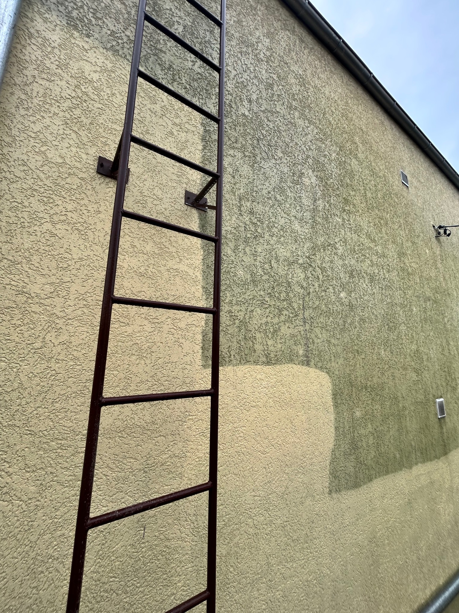 Elewacja budynku z częściowo pomalowaną ścianą w dwóch odcieniach beżu, z metalową drabiną przymocowaną do ściany. Widoczny fragment rynny.