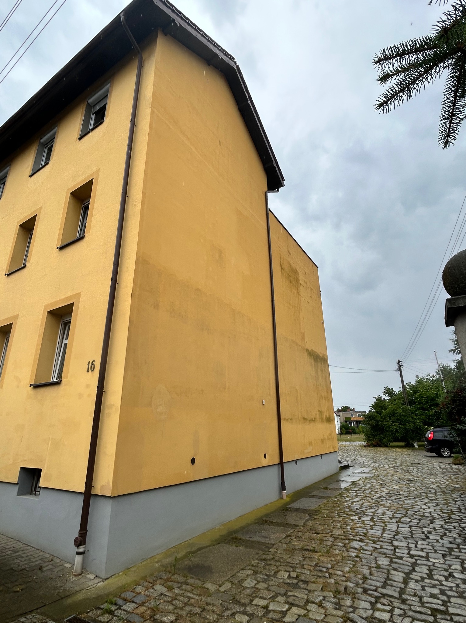 Żółta elewacja budynku z numerem 16 na boku, widoczny fragment dachu i rynna, obok brukowana ulica. Pochmurne niebo w tle.