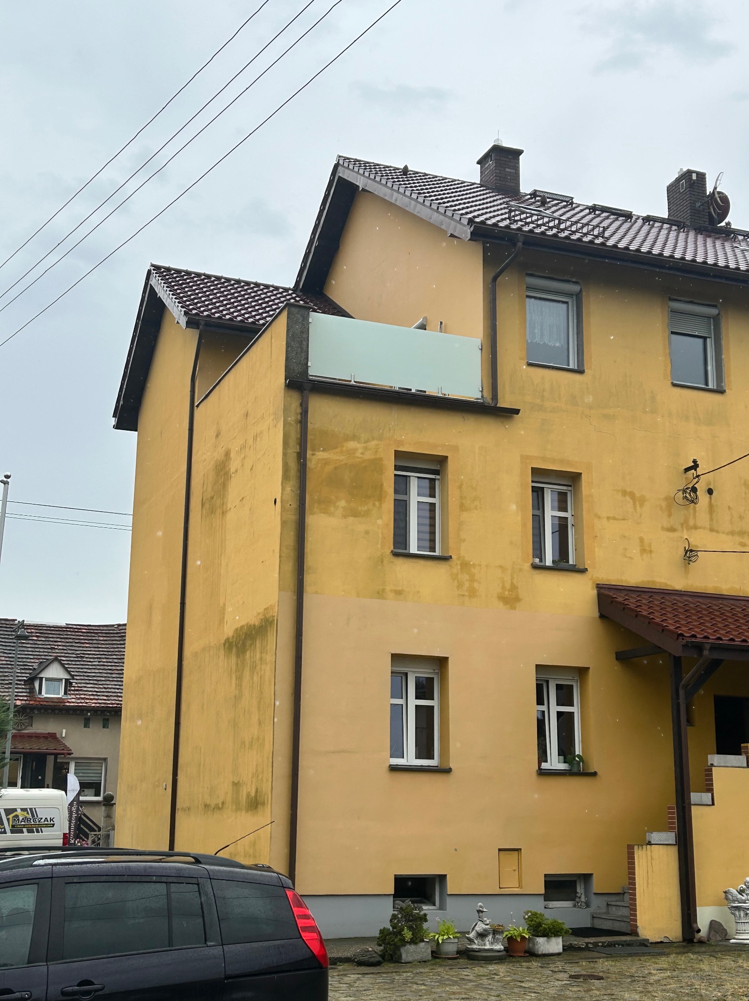 Żółta, dwukondygnacyjna kamienica z balkonem w Michałowie Drugim, z widocznymi śladami zabrudzeń na elewacji i fragmentem zaparkowanego samochodu na pierwszym planie.