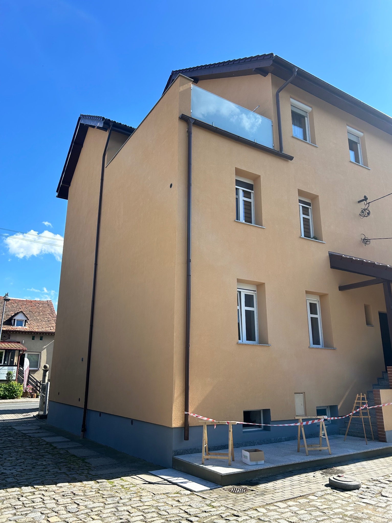 Ocieplona, pomalowana na beżowo elewacja budynku z balkonem i brązowymi rynnami. Na dole rozstawione barierki i rusztowania.