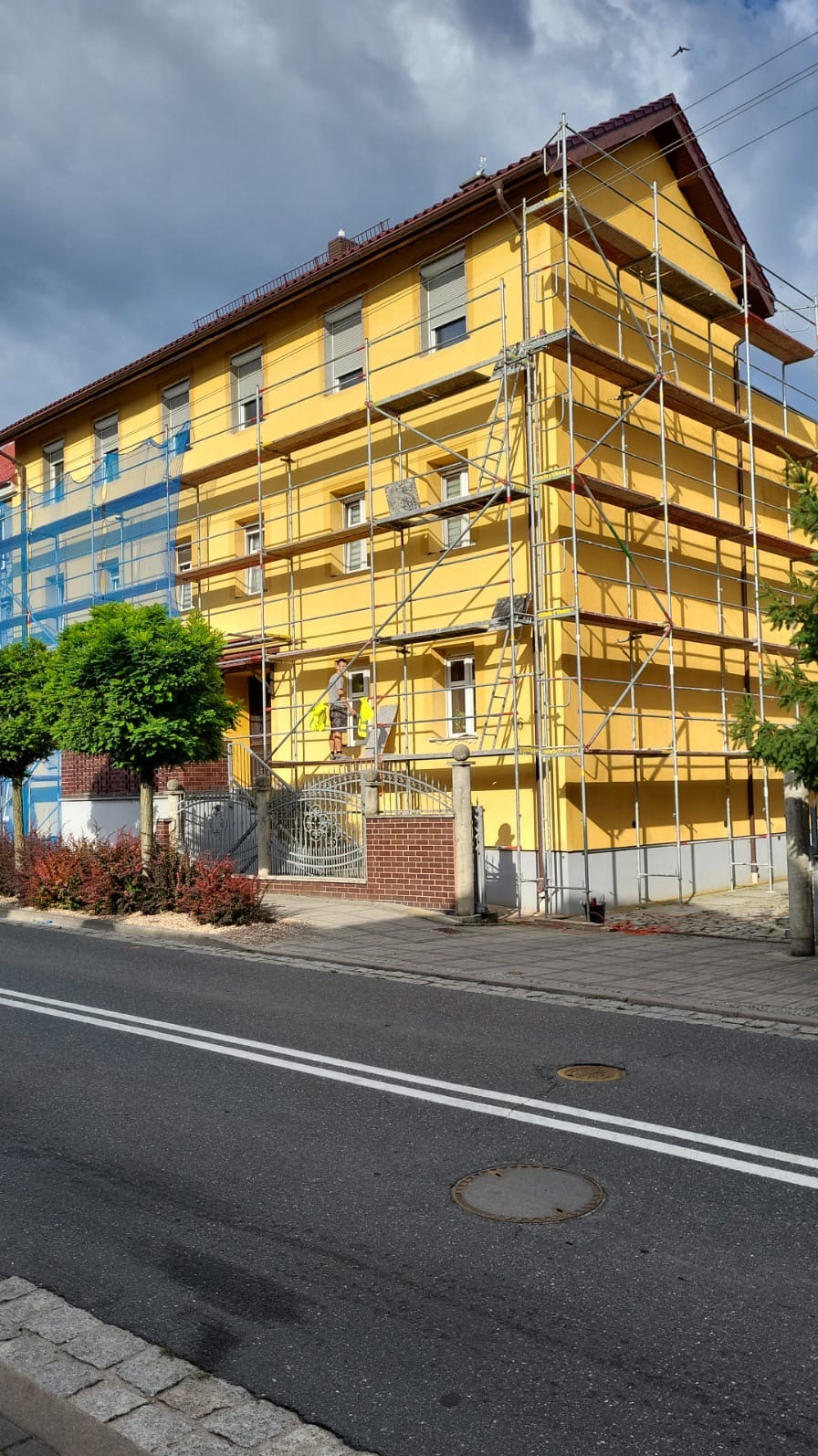 Żółty dom w rusztowaniach, pracownik w kamizelce maluje elewację. Ujęcie z dołu, widoczne drzewa i ulica. Słoneczny dzień, minimalne zachmurzenie.