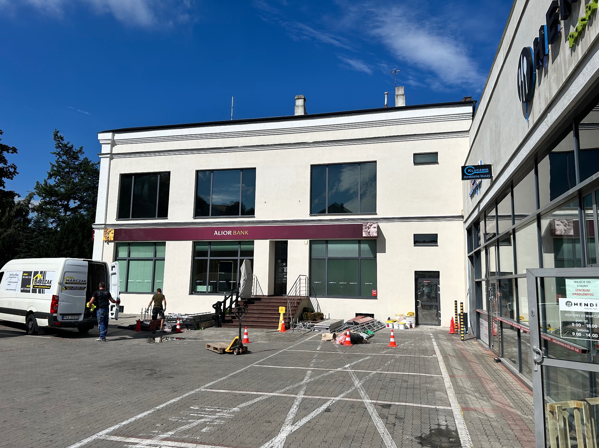 Przygotowania do prac elewacyjnych w Michałowie Drugim. Rozstawione rusztowania i zabezpieczenia przed budynkiem Alior Banku i Hendi. Pracownicy w trakcie organizacji.