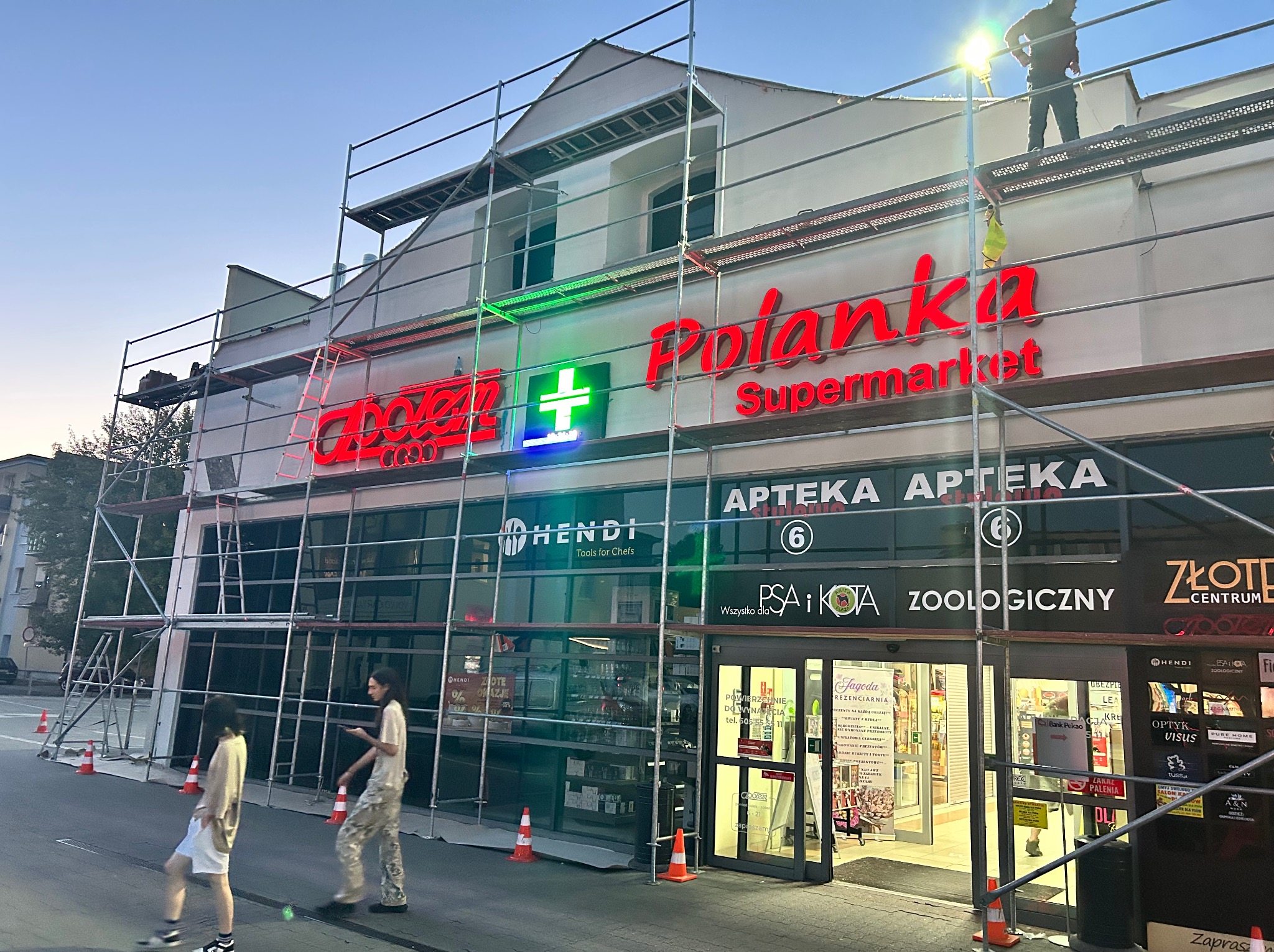 Elewacja budynku sklepu w rusztowaniach przed malowaniem, widoczne neonowe szyldy i osoby przechodzące obok. Prace elewacyjne w toku, pomarańczowe pachołki ostrzegawcze.