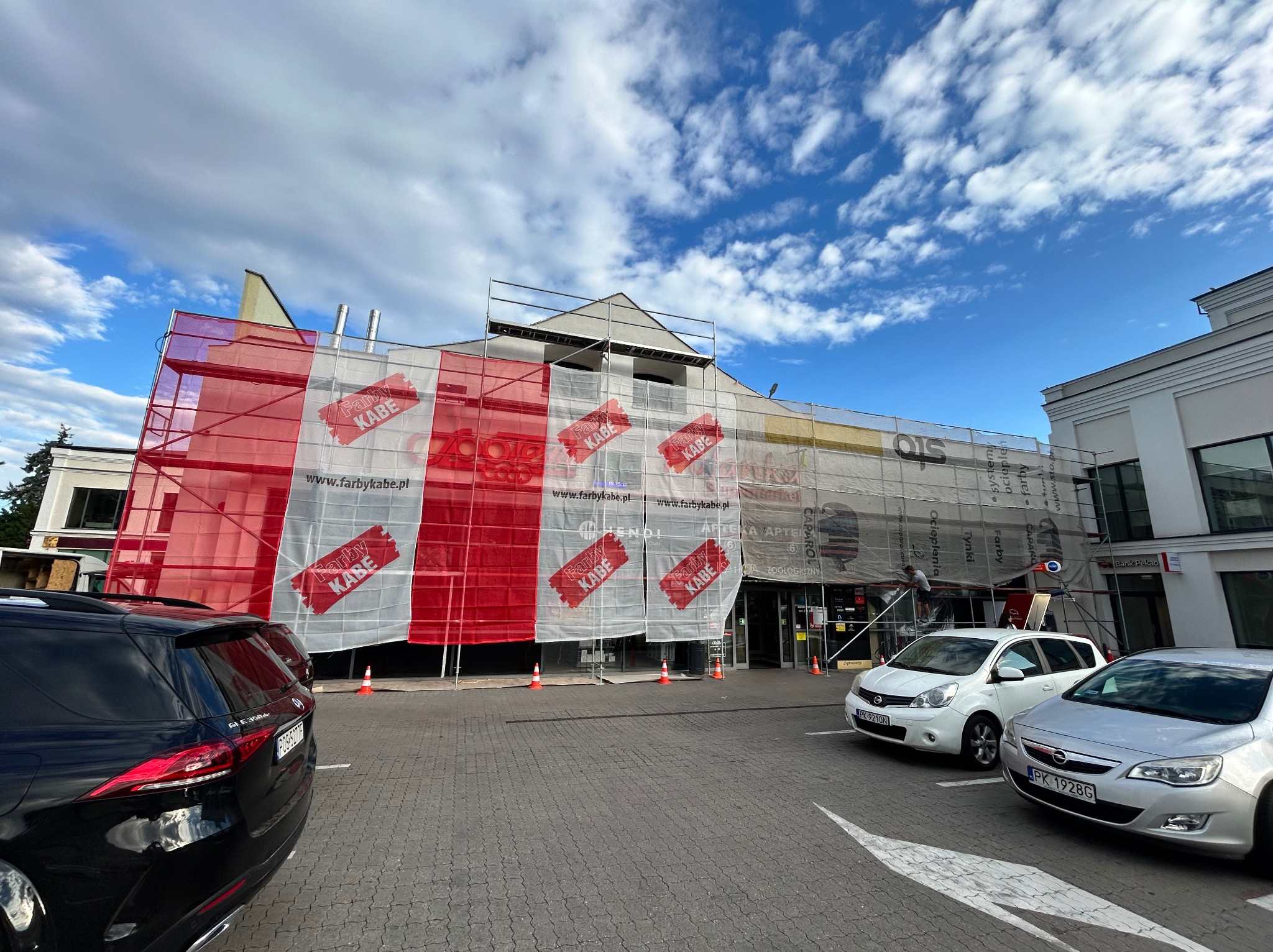 Budynek w rusztowaniach osłonięty siatką z reklamą Farby KABE. Widoczne zaparkowane samochody i elementy otoczenia miejskiego.