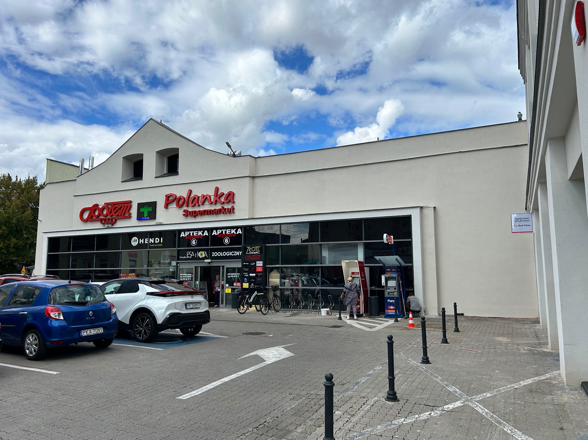 Budynek supermarketu z białymi elewacjami i czerwonym napisem 'Polanka'. Widoczne zaparkowane samochody i osoba przy bankomacie. Błękitne niebo z chmurami w tle.