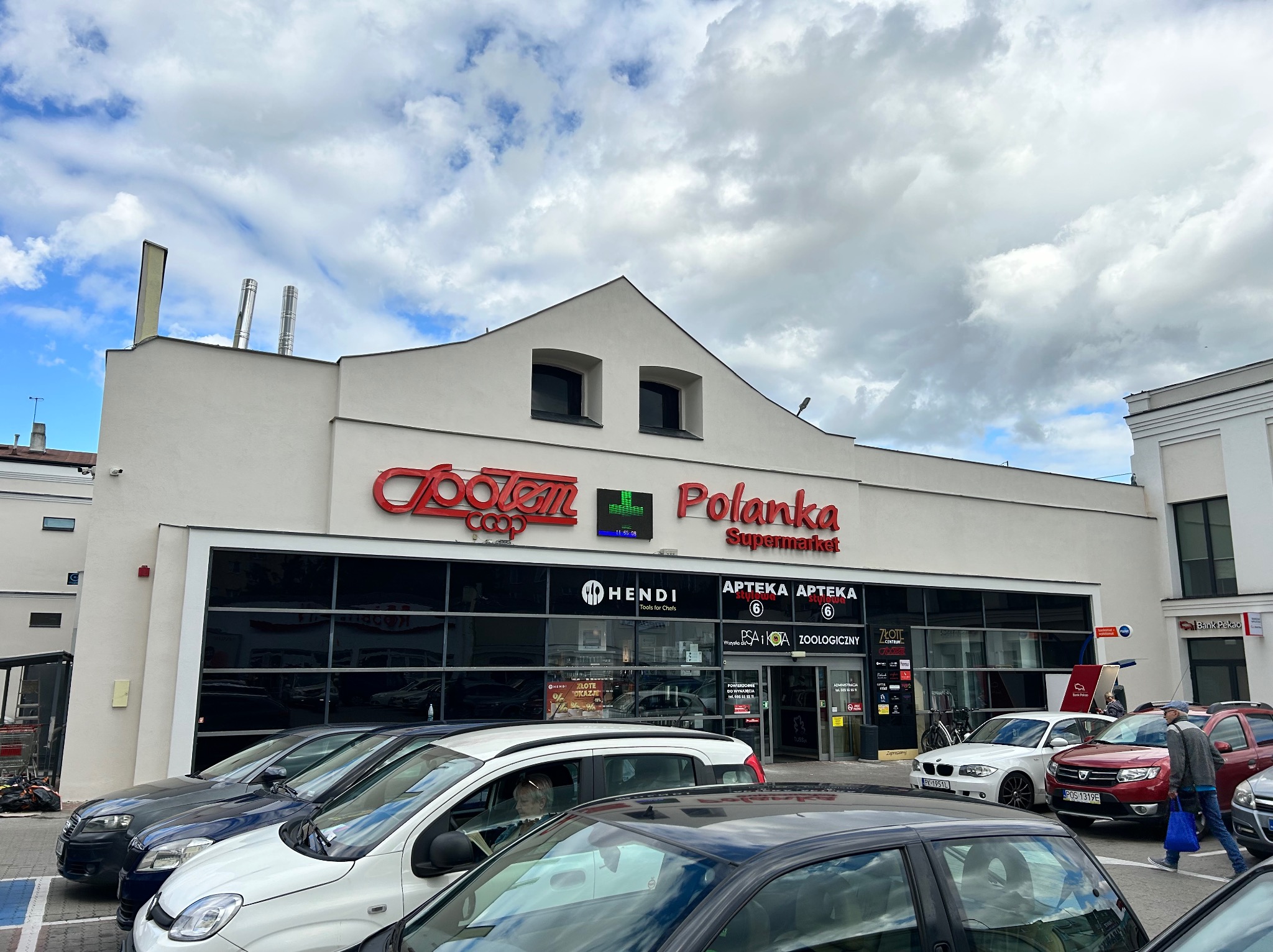 Biała elewacja budynku sklepu 'Polanka' z czerwonym logo 'Społem Coop'. Widoczne zaparkowane samochody i fragment chodnika z przechodniem.