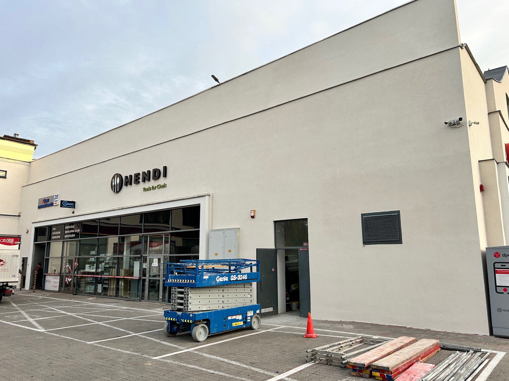 Jasna elewacja budynku z logo firmy Hendi, widoczny podnośnik koszowy i rusztowania. Świeżo położony tynk strukturalny. Prace wykończeniowe.