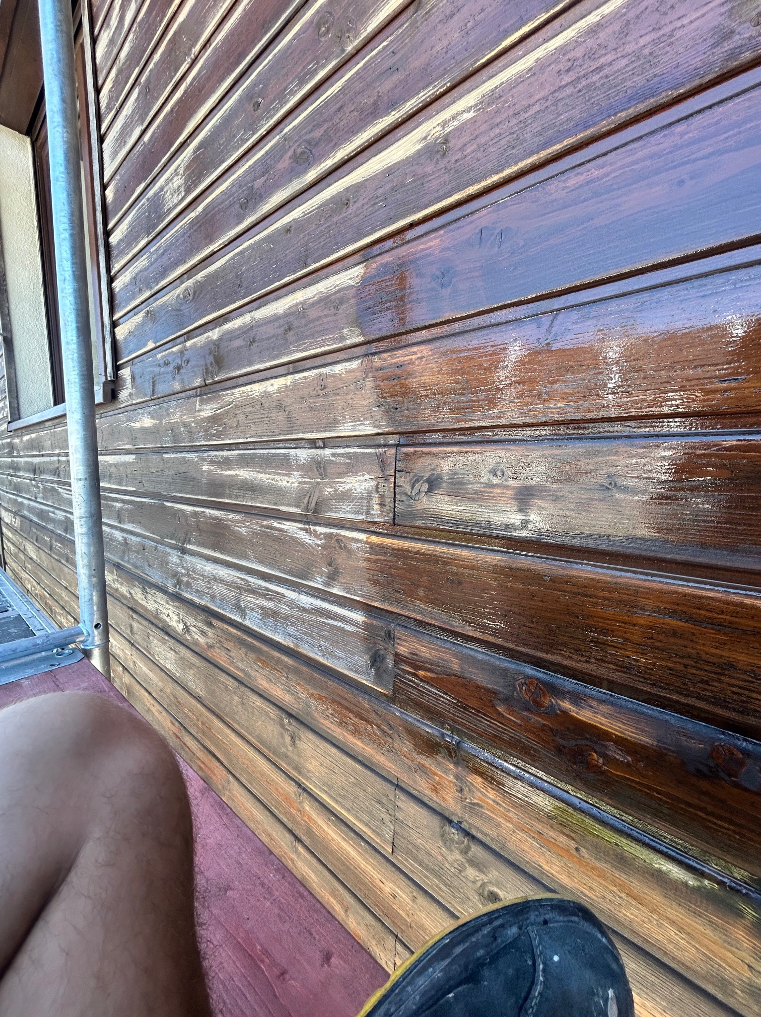 Świeżo pomalowana, błyszcząca, brązowa drewniana elewacja typu siding. Widoczne fragmenty rusztowania i buta. Ujęcie z dołu, podkreślające strukturę drewna.