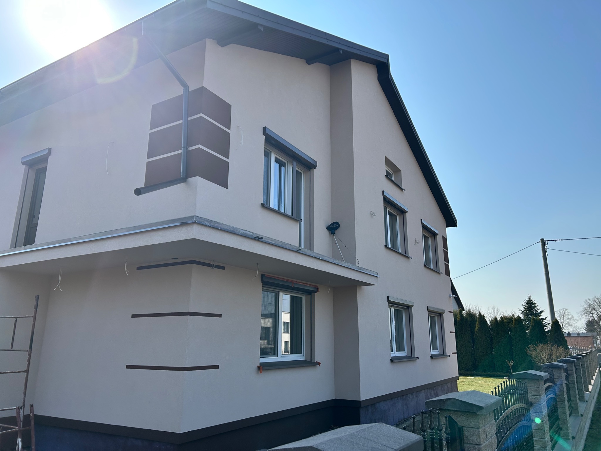 Jasna elewacja domu z ciemnymi akcentami dekoracyjnymi, widoczne okna z roletami, fragment drabiny przy ścianie. Słoneczny dzień, błękitne niebo w tle.