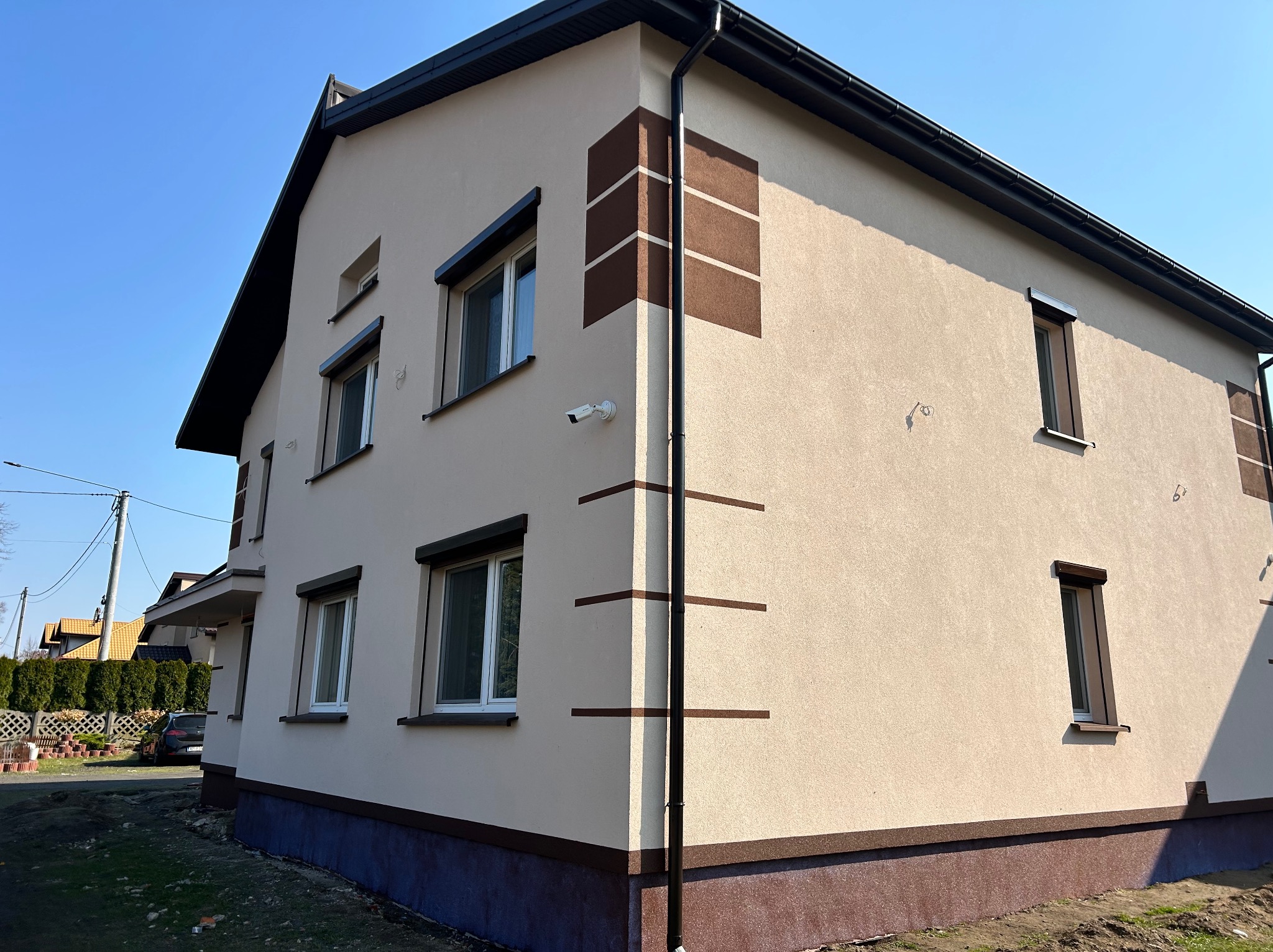 Dwukondygnacyjny dom z beżową elewacją i brązowymi akcentami dekoracyjnymi. Widoczne okna z roletami, rynna i kamera. Uwagę przykuwa narożne zdobienie w postaci pasów.