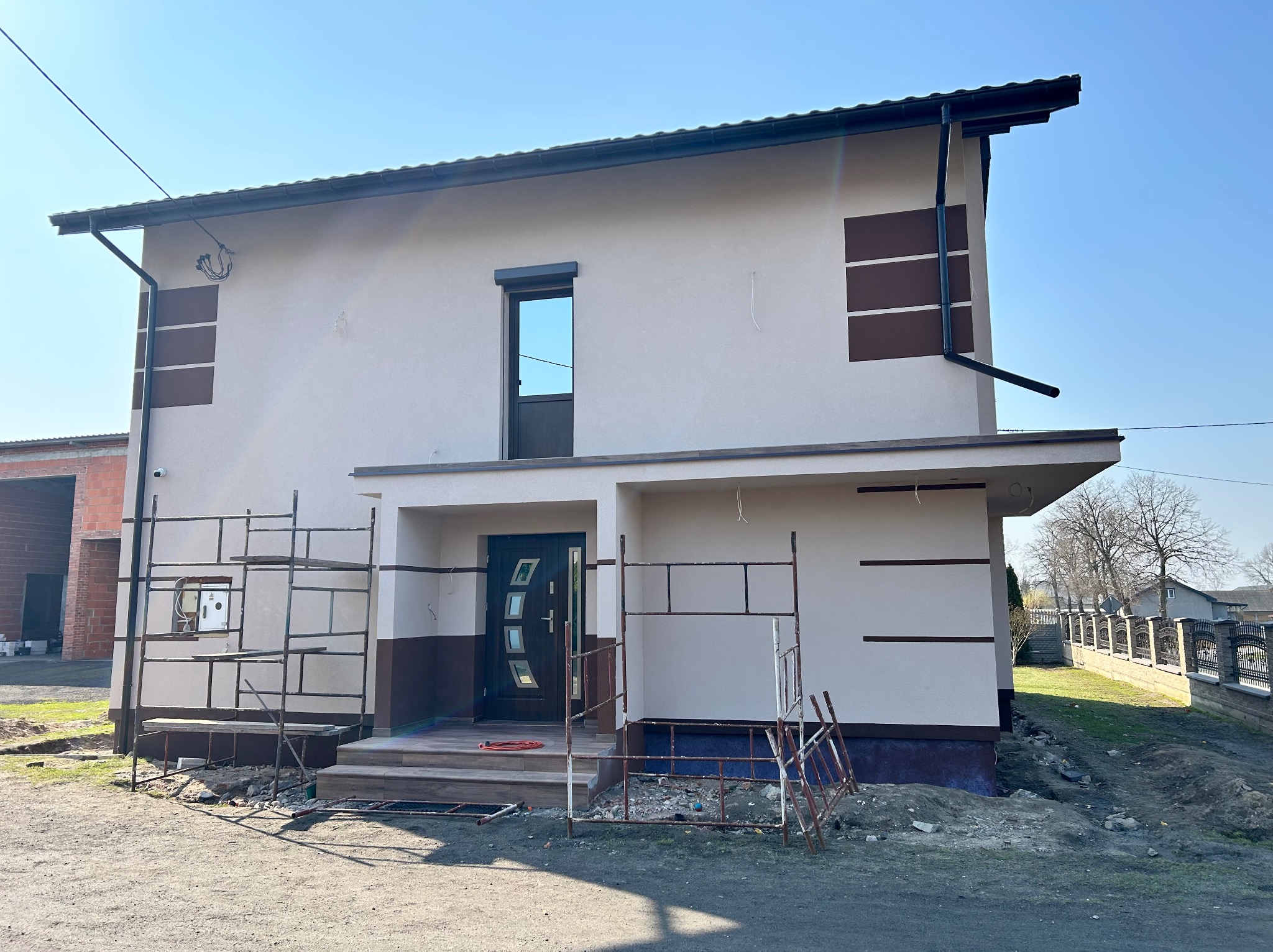 Dwukondygnacyjny dom z jasną elewacją i ciemnymi akcentami, w trakcie prac wykończeniowych. Widoczne rusztowania i teren wokół domu w trakcie zagospodarowania.