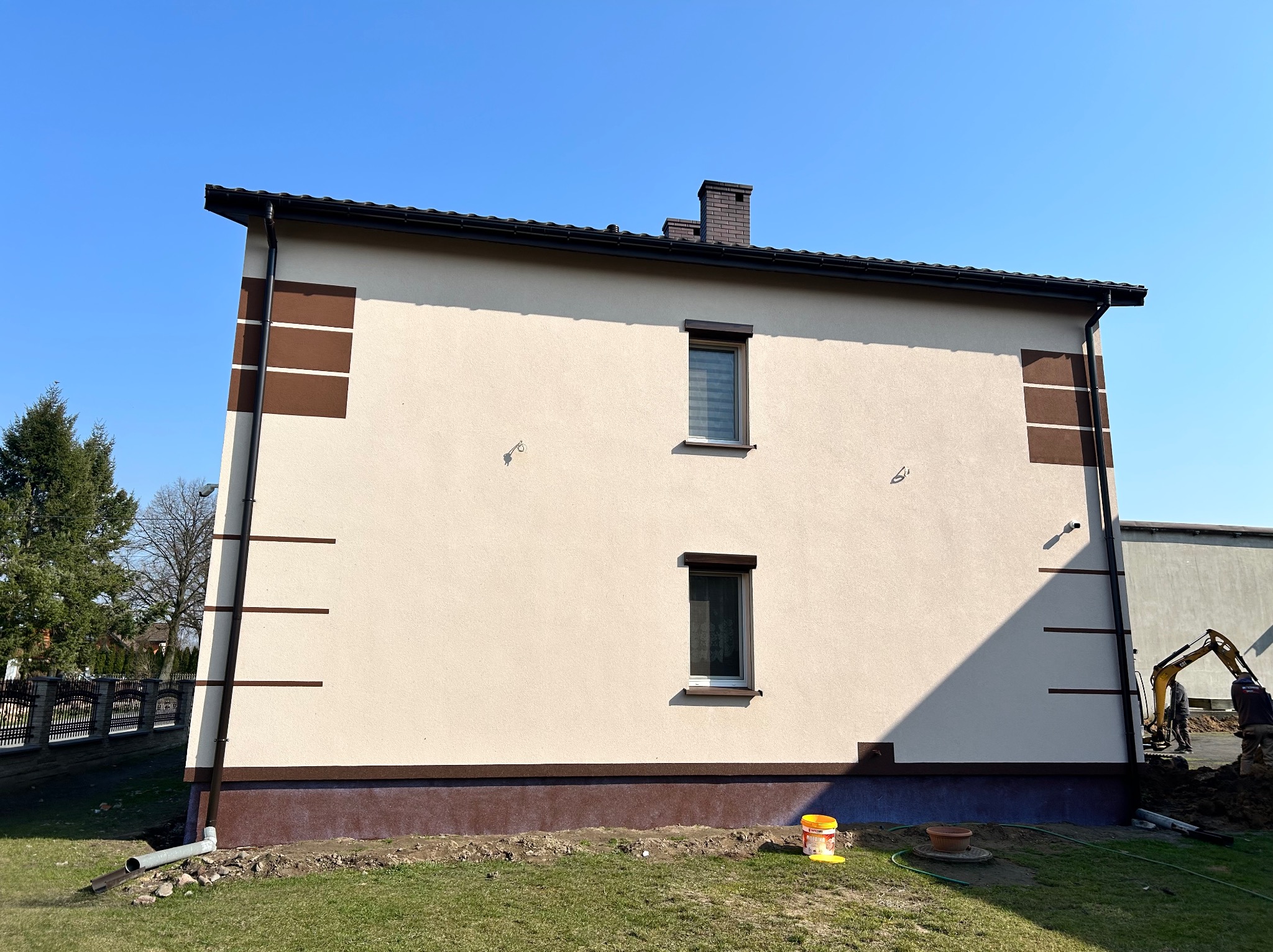Elewacja domu po renowacji z brązowymi akcentami, widoczne okna z roletami i fragment dachu. Na trawniku stoi wiadro z farbą. W tle prace ziemne.