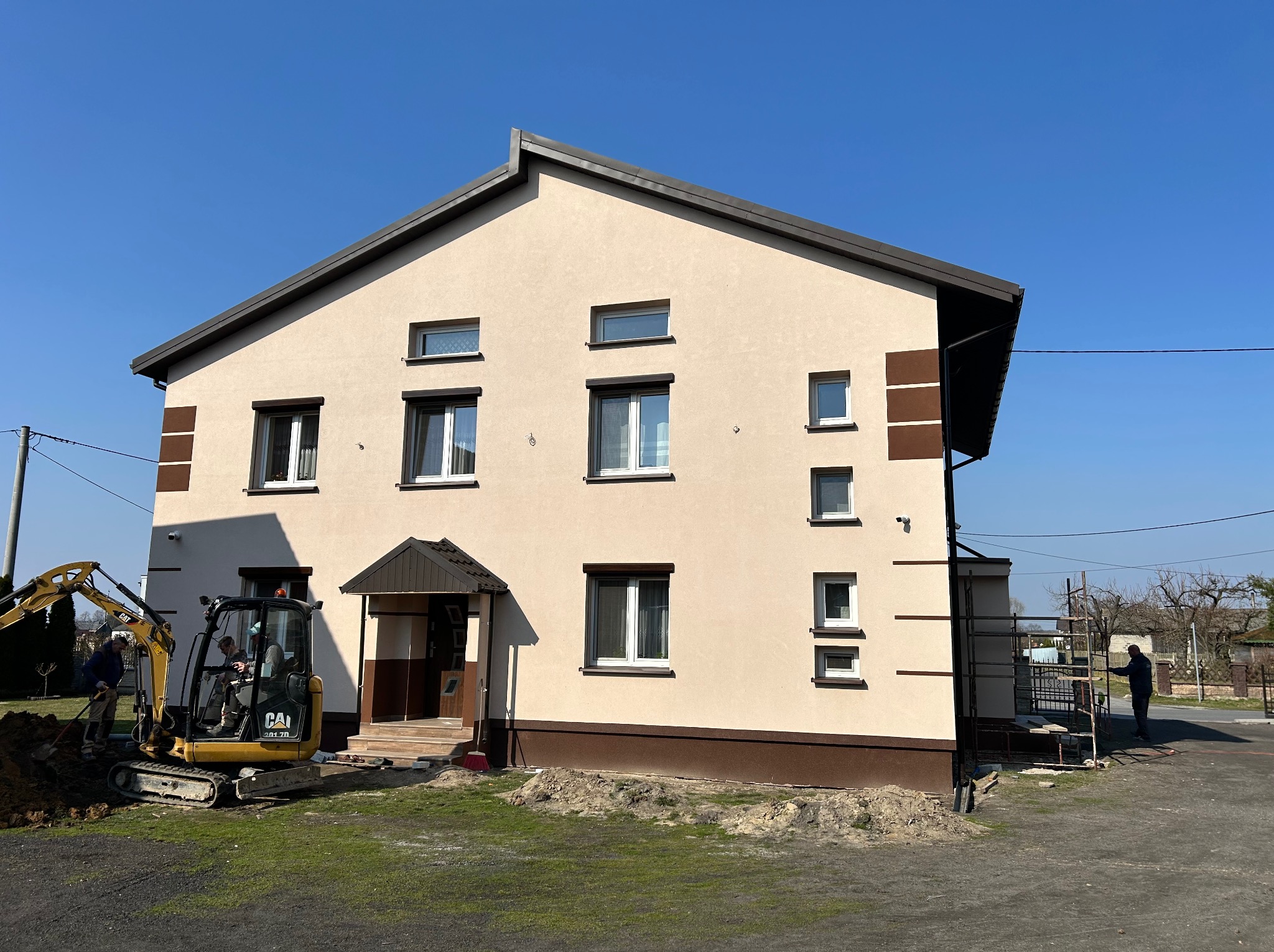 Dwukondygnacyjny dom z beżową elewacją i brązowymi akcentami. Na pierwszym planie minikoparka i pracownicy, z boku rusztowanie. Błękitne niebo w tle.