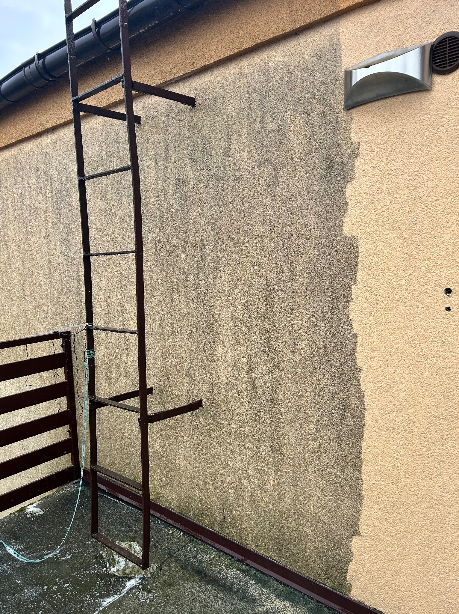 Fragment elewacji z drabiną, ukazujący różnicę w czystości przed i po myciu. Widoczne zabrudzenia na części ściany, kontrastujące z czystą, jasną powierzchnią.