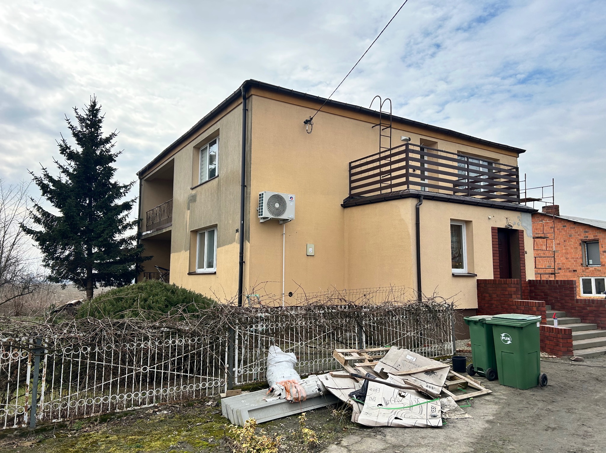 Dwupiętrowy dom z beżową elewacją, balkonem i klimatyzacją. Widoczne ślady prac remontowych, rusztowanie i śmieci przy ogrodzeniu. Pochmurne niebo w tle.