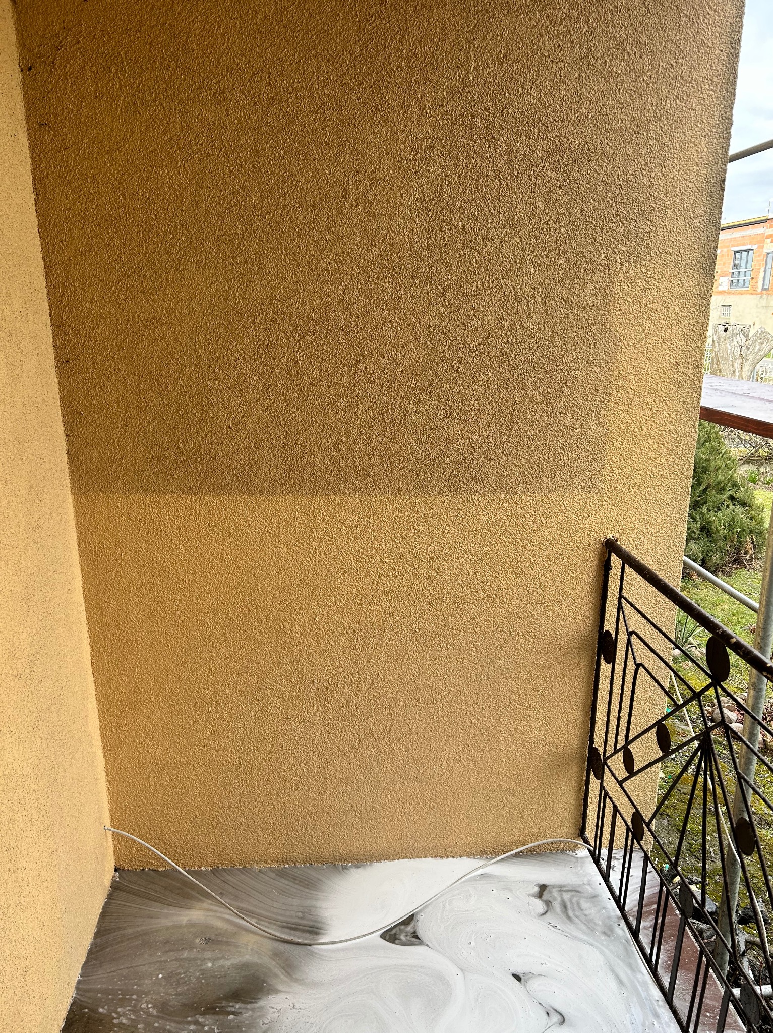 Fragment elewacji budynku z widoczną różnicą w kolorze po czyszczeniu. Na balkonie woda z detergentem. Metalowa balustrada balkonu z geometrycznym wzorem.