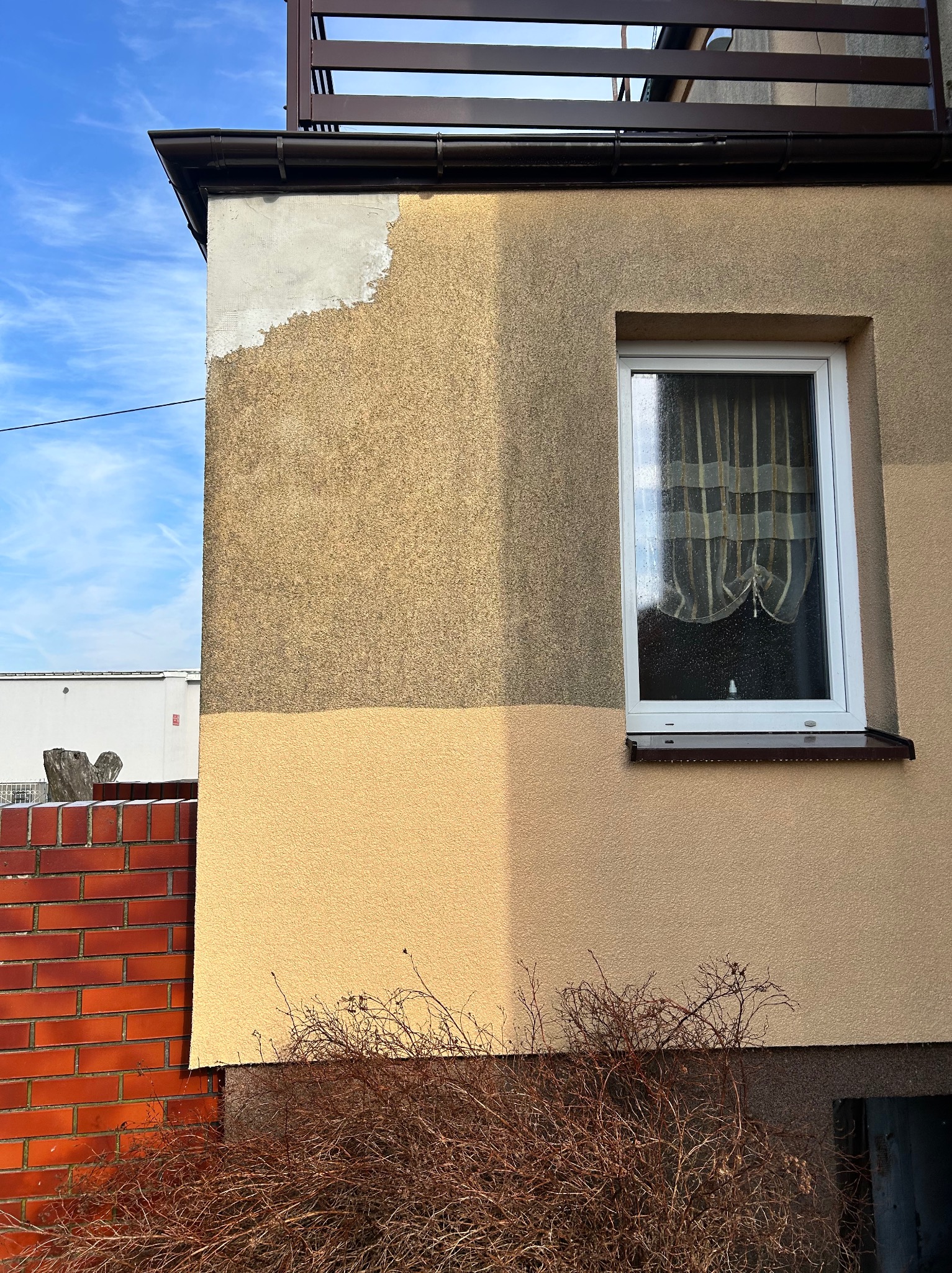 Fragment elewacji budynku w trakcie prac: część ściany otynkowana, część pomalowana na dwa odcienie beżu. Widoczne okno z zasłonką i ceglany murek.