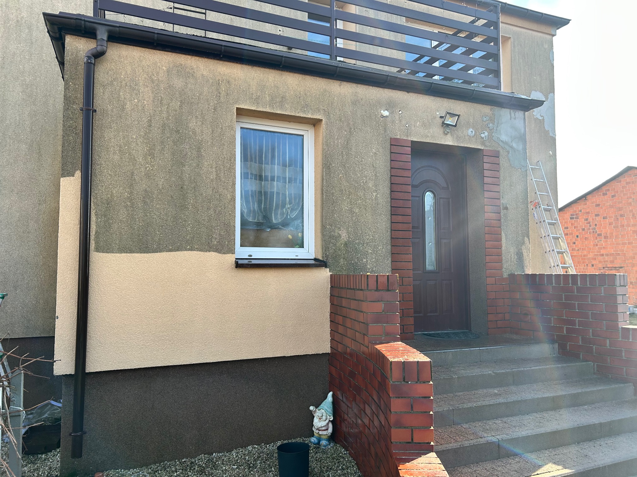 Dwukolorowa elewacja domu z oknem, ceglanym wejściem i drabina oparta o ścianę. Część ściany wymaga naprawy. Widoczny krasnal ogrodowy.