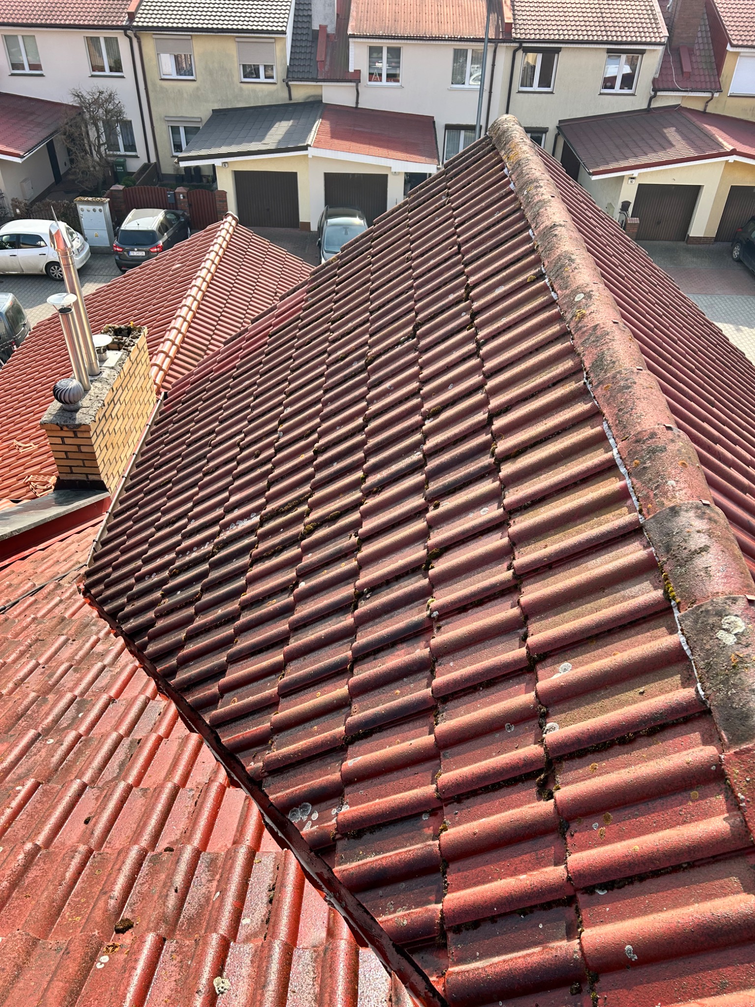 Czerwony dach z dachówki karpiówki, widoczny z góry, z mchem i zabrudzeniami. W tle zabudowa jednorodzinna z garażami. Słoneczny dzień.