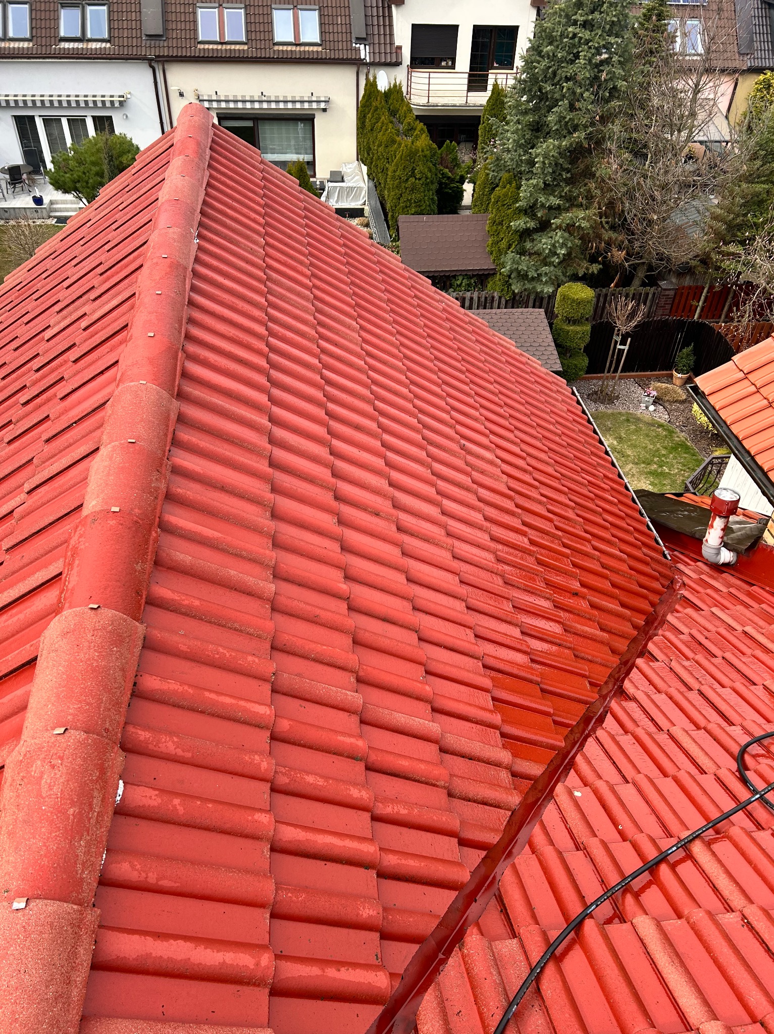 Czerwona dachówka ceramiczna po czyszczeniu, widoczne ślady wilgoci. W tle zabudowa jednorodzinna i zieleń. Ujęcie z góry, lekki kąt.