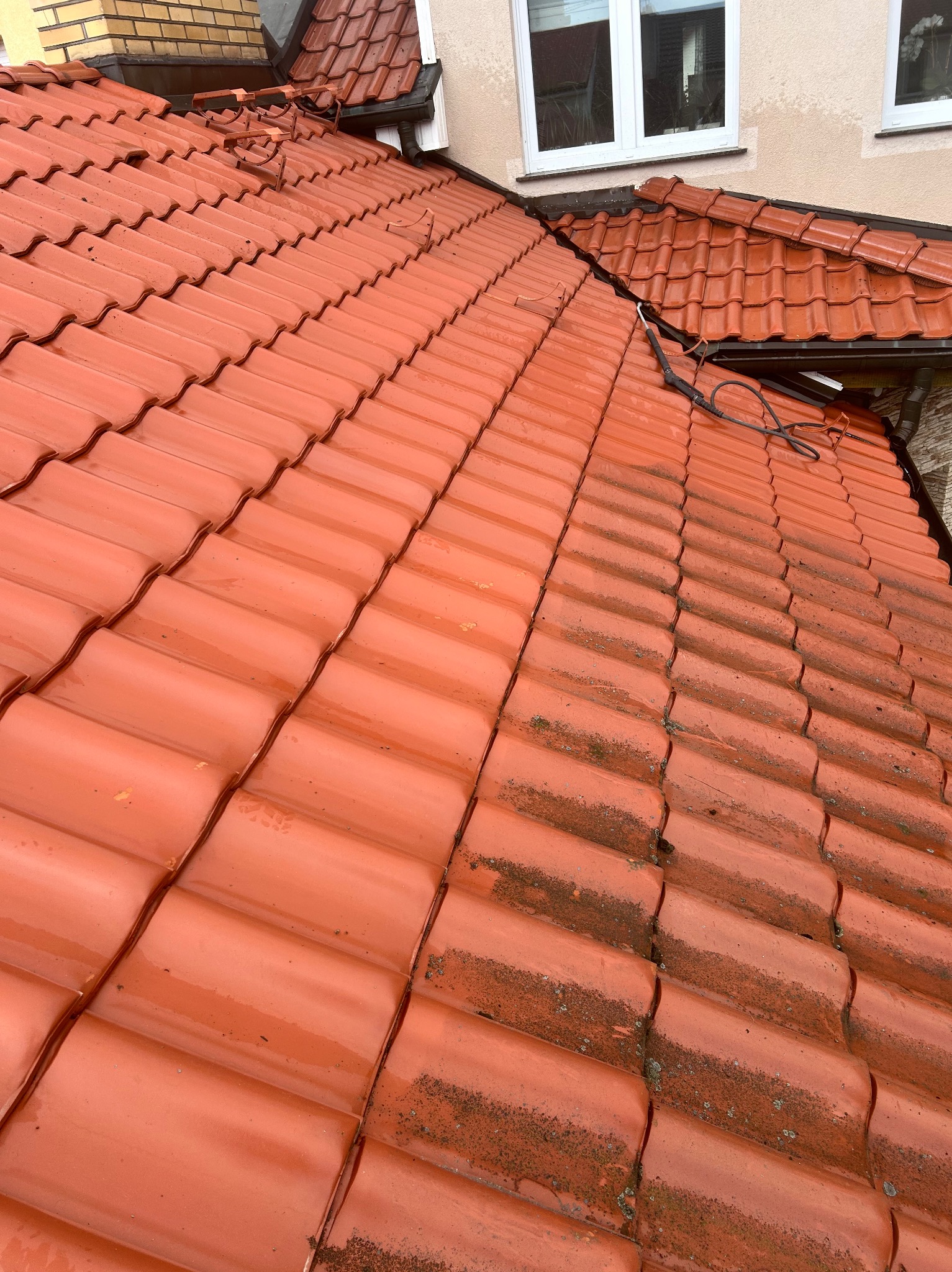 Częściowo umyty, mokry dach z czerwonej dachówki karpiówki. Widoczny kontrast czystej i zabrudzonej powierzchni. Ujęcie z góry, lekki skos.