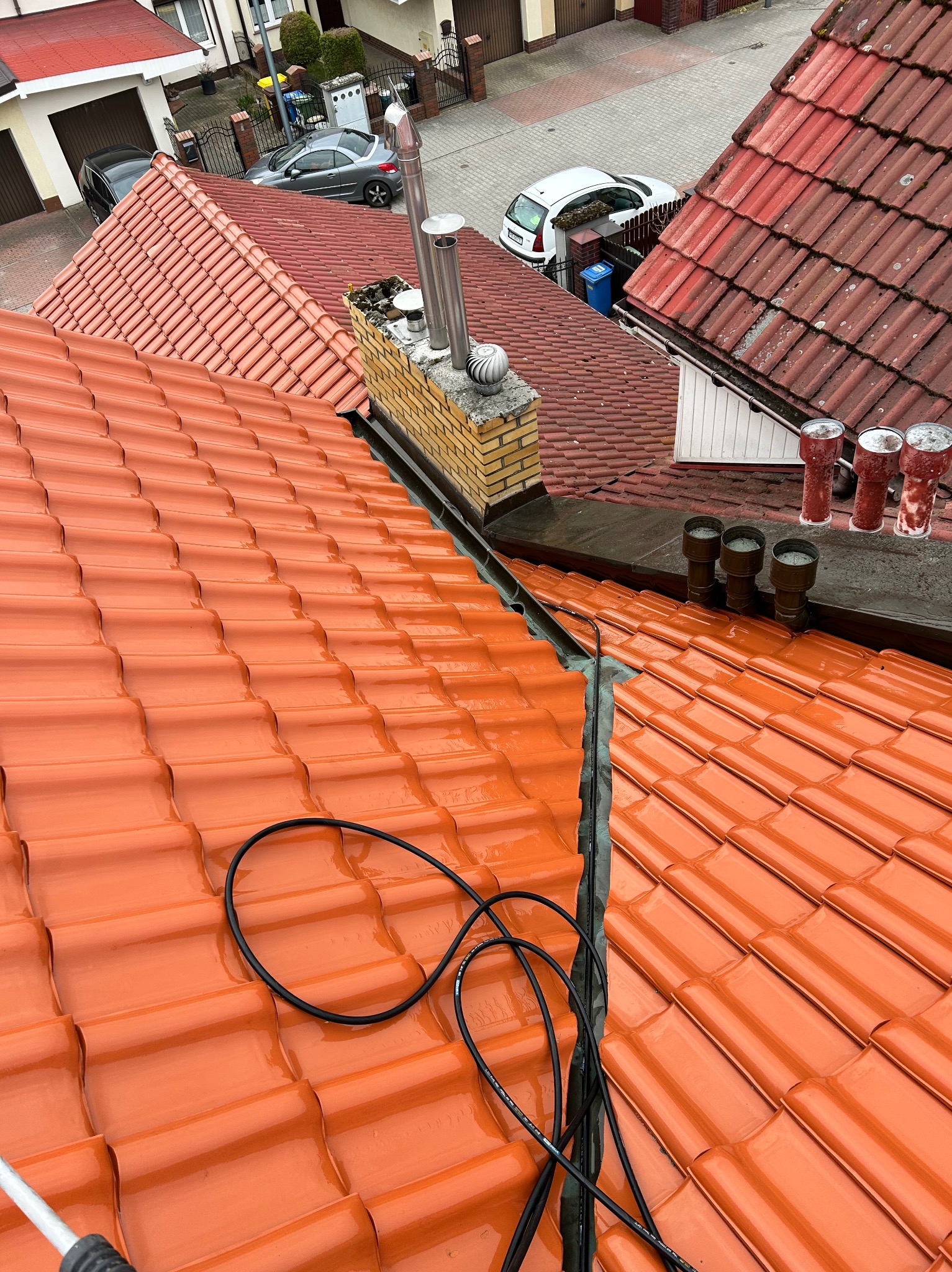 Błyszcząca, pomarańczowa dachówka ceramiczna po myciu, widok z góry. W tle komin z cegły i fragmenty innych dachów. Widoczne przewody i lanca myjki ciśnieniowej.