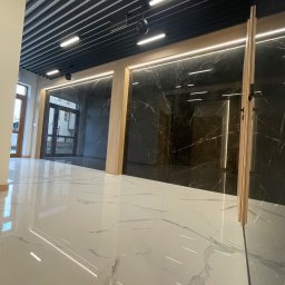 PerfectHouse Wykończenia Wnętrz Kamil Pazdan - Wykończone wnętrze z błyszczącą białą marmurową podłogą, czarnymi panelami ściennymi z marmurowym wzorem i drewnianymi akcentami, oświetlone nowoczesnymi lampami sufitowymi.