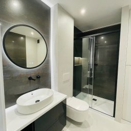 PerfectHouse Wykończenia Wnętrz Kamil Pazdan - Nowoczesna łazienka z owalną umywalką nablatową, okrągłym lustrem z podświetleniem LED, czarną armaturą i kabiną prysznicową z ciemnymi szybami.