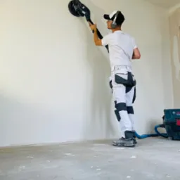 Pracownik w okularach ochronnych i słuchawkach szlifuje ścianę za pomocą szlifierki oscylacyjnej podłączonej do odkurzacza przemysłowego, widoczne białe spodnie robocze z kieszeniami.