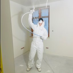 PerfectHouse Wykończenia Wnętrz Kamil Pazdan - Pracownik w białym kombinezonie ochronnym i masce maluje ściany natryskowo w jasnym pomieszczeniu, okno zabezpieczone folią, widoczne gniazdka elektryczne w ścianie.