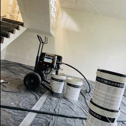 PerfectHouse Wykończenia Wnętrz Kamil Pazdan - Agregat malarski stoi na folii ochronnej w pomieszczeniu ze schodami, obok ustawione wiadra z farbą, ściany przygotowane do malowania.