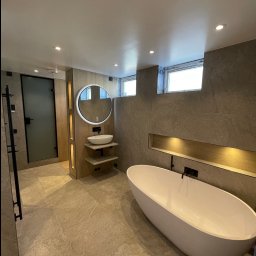 PerfectHouse Wykończenia Wnętrz Kamil Pazdan - Nowoczesna łazienka z wolnostojącą wanną, okrągłym lustrem z podświetleniem LED, umywalką nablatową na drewnianych półkach i szklaną kabiną prysznicową z czarnymi elementami.