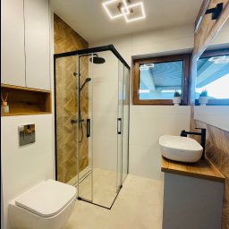 PerfectHouse Wykończenia Wnętrz Kamil Pazdan - Nowoczesna łazienka z kabiną prysznicową w czarnej ramie, podwieszaną toaletą, umywalką nablatową na drewnianym blacie i geometrycznym oświetleniem sufitowym.