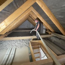 PerfectHouse Wykończenia Wnętrz Kamil Pazdan - Pracownik w masce aplikuje izolację z wełny mineralnej na poddaszu, widoczne drewniane krokwie i rury do transportu materiału izolacyjnego.
