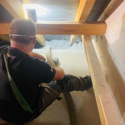 PerfectHouse Wykończenia Wnętrz Kamil Pazdan - Pracownik w czarnej koszulce z logo, w okularach ochronnych, aplikuje izolację z włókien w poddaszu, używając węża i specjalistycznego sprzętu. Widoczne drewniane belki konstrukcyjne i częściowo...