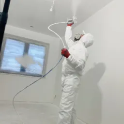 Pracownik w kombinezonie ochronnym i czerwonych rękawicach maluje sufit agregatem natryskowym; okno zabezpieczone folią malarską.