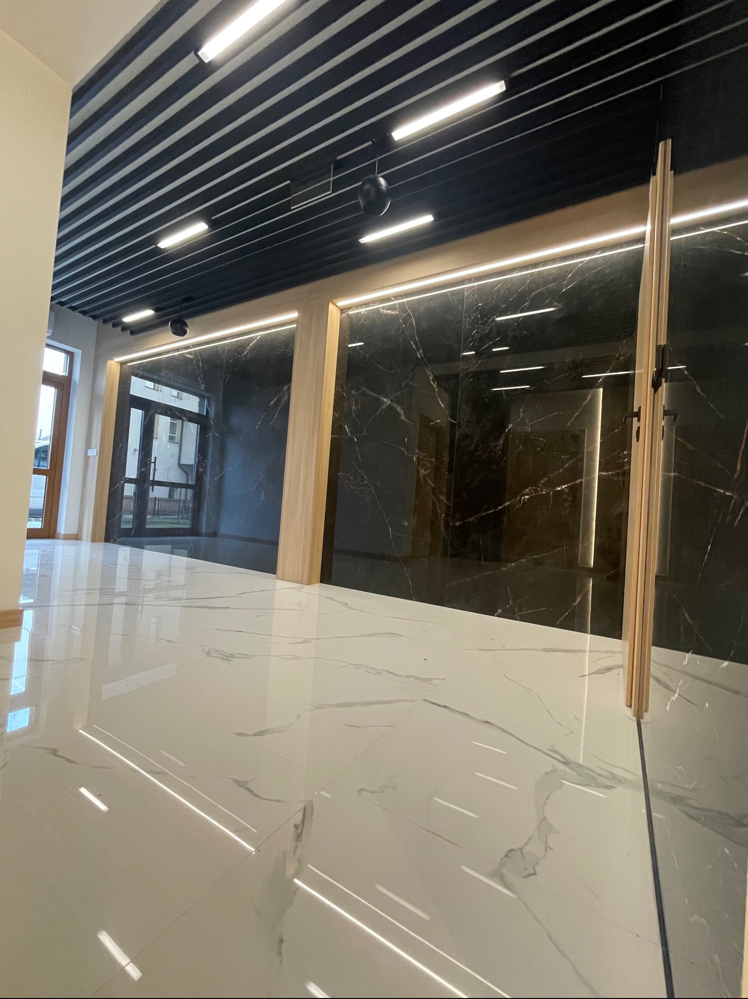 Wykończone wnętrze z błyszczącą białą marmurową podłogą, czarnymi panelami ściennymi z marmurowym wzorem i drewnianymi akcentami, oświetlone nowoczesnymi lampami sufitowymi.