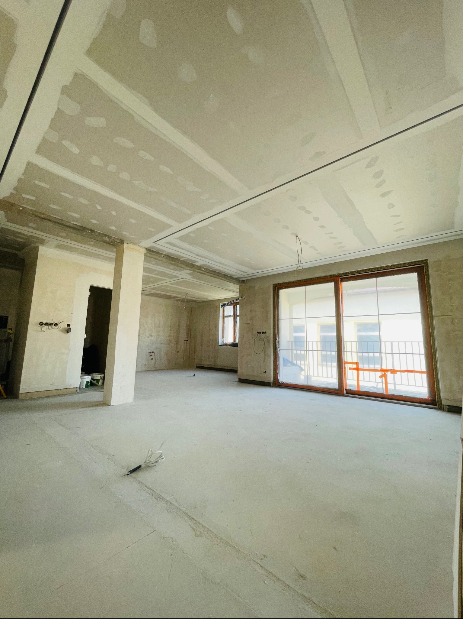 Wnętrze w trakcie remontu: sufit z płyt kartonowo-gipsowych z widocznymi łączeniami, ściany przygotowane pod malowanie, duże okno przesuwne z widokiem na balkon, szpachla na podłodze, widoczne...