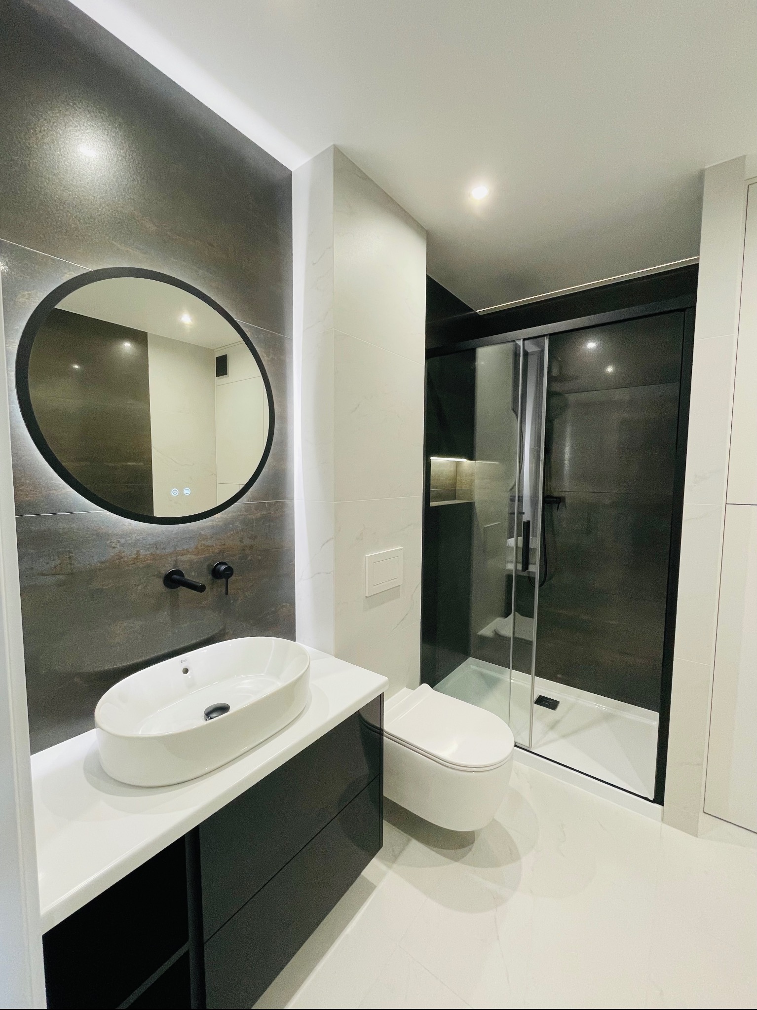 Nowoczesna łazienka z owalną umywalką nablatową, okrągłym lustrem z podświetleniem LED, czarną armaturą i kabiną prysznicową z ciemnymi szybami.