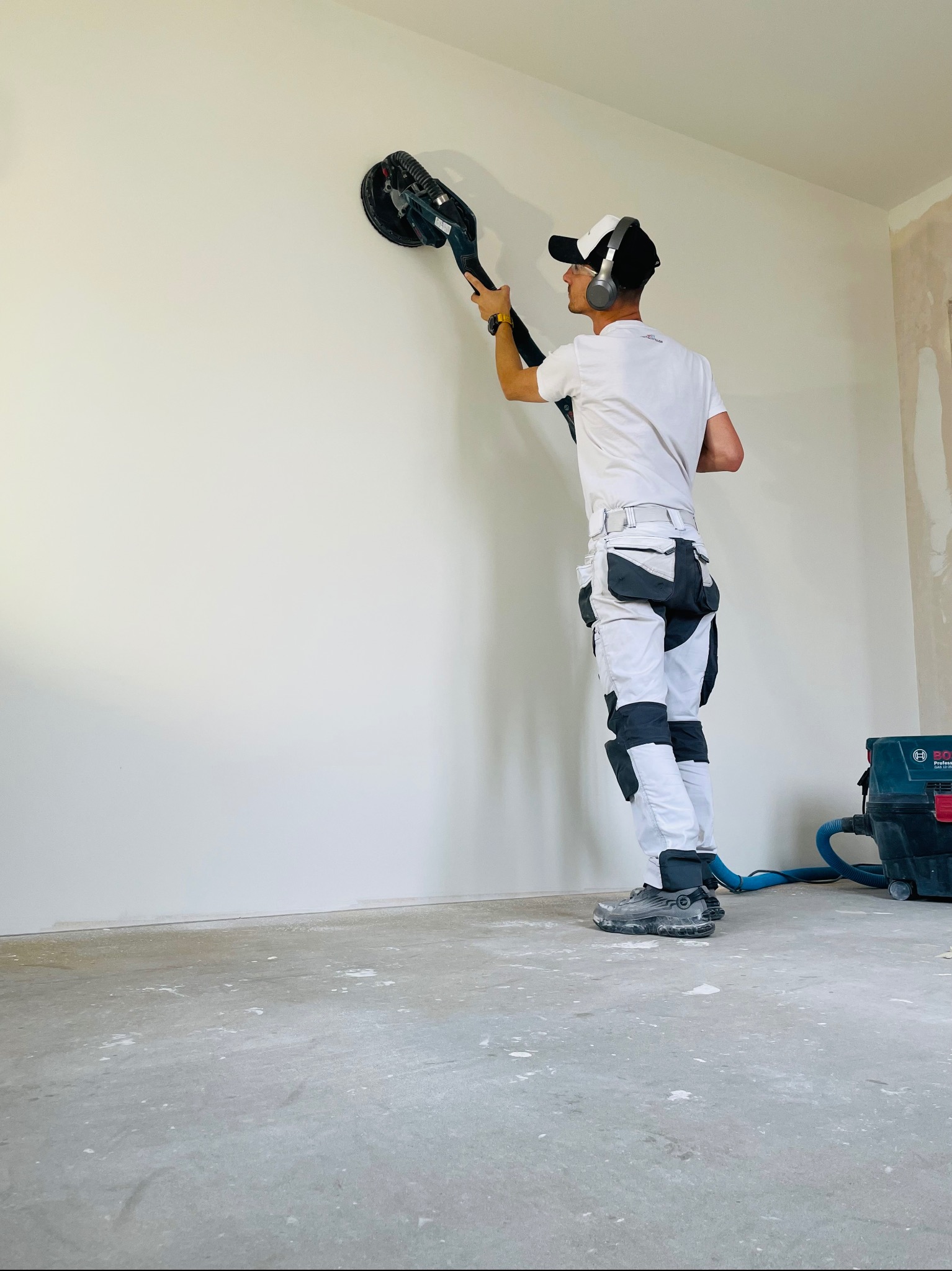 Pracownik w okularach ochronnych i słuchawkach szlifuje ścianę za pomocą szlifierki oscylacyjnej podłączonej do odkurzacza przemysłowego, widoczne białe spodnie robocze z kieszeniami.