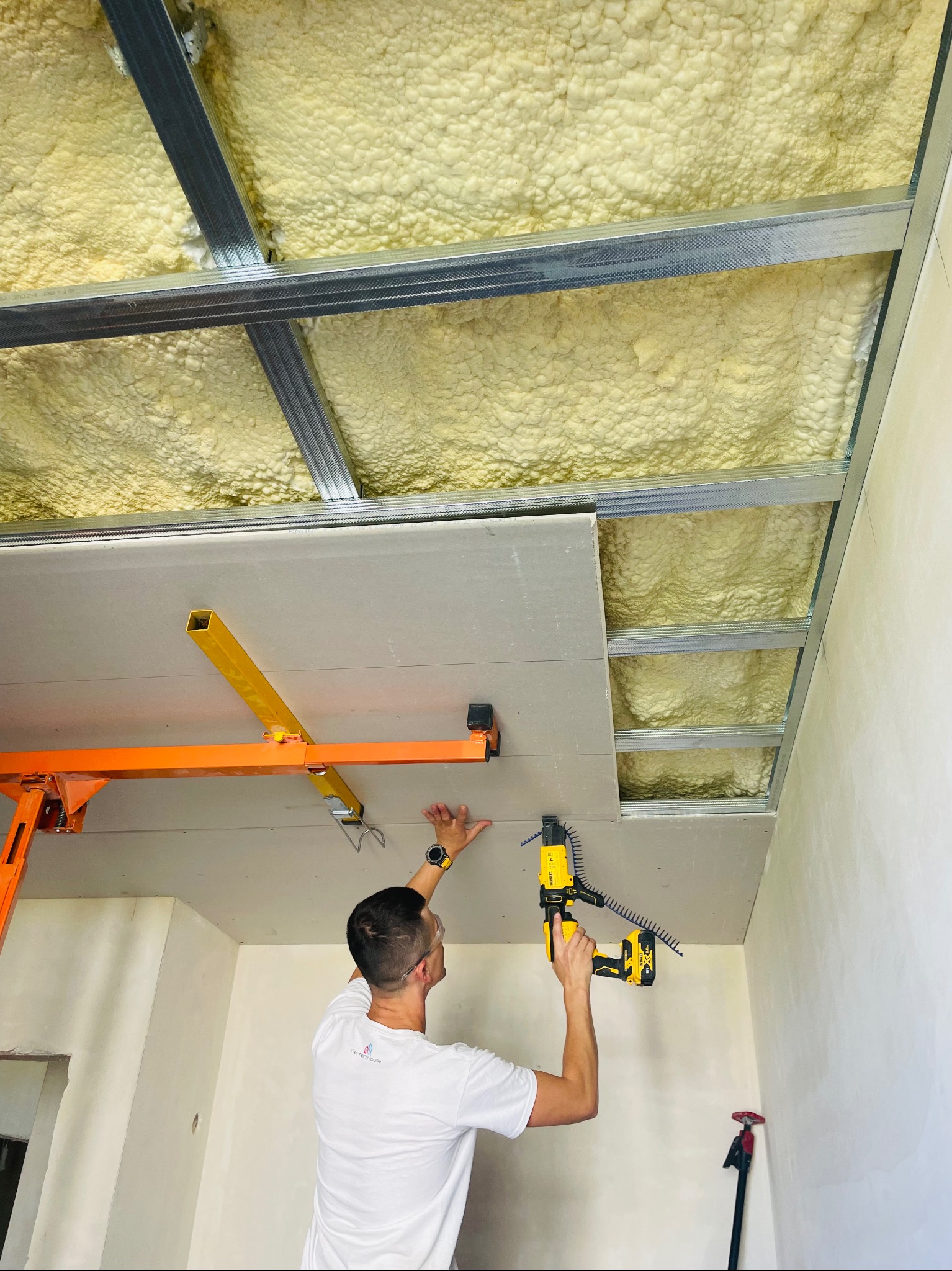 Pracownik montuje płytę karton gips na metalowym stelażu pod sufitem, widoczna warstwa ocieplenia poddasza, używa wkrętarki automatycznej DeWalt i podnośnika do płyt.