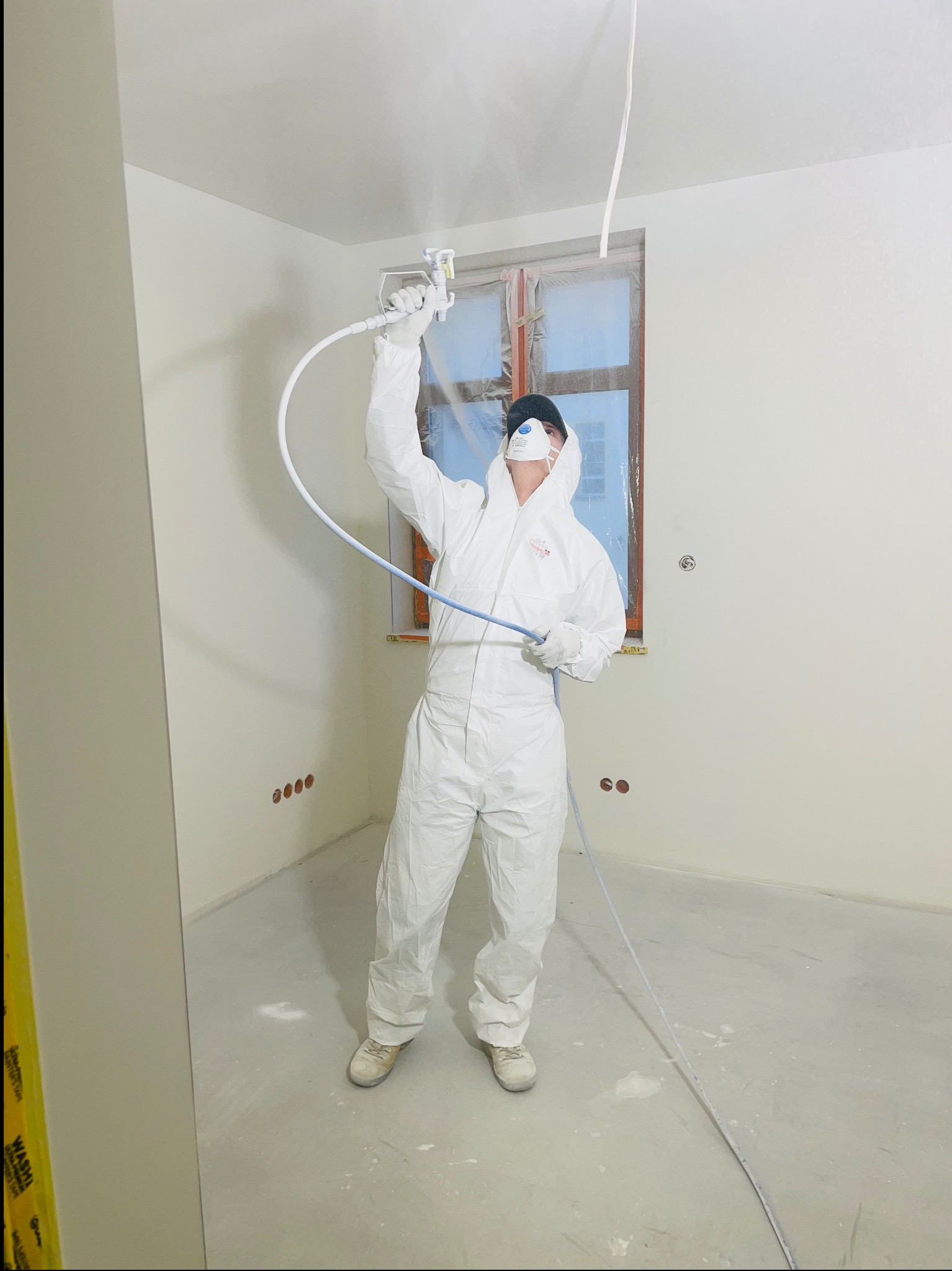 Pracownik w białym kombinezonie ochronnym i masce maluje ściany natryskowo w jasnym pomieszczeniu, okno zabezpieczone folią, widoczne gniazdka elektryczne w ścianie.