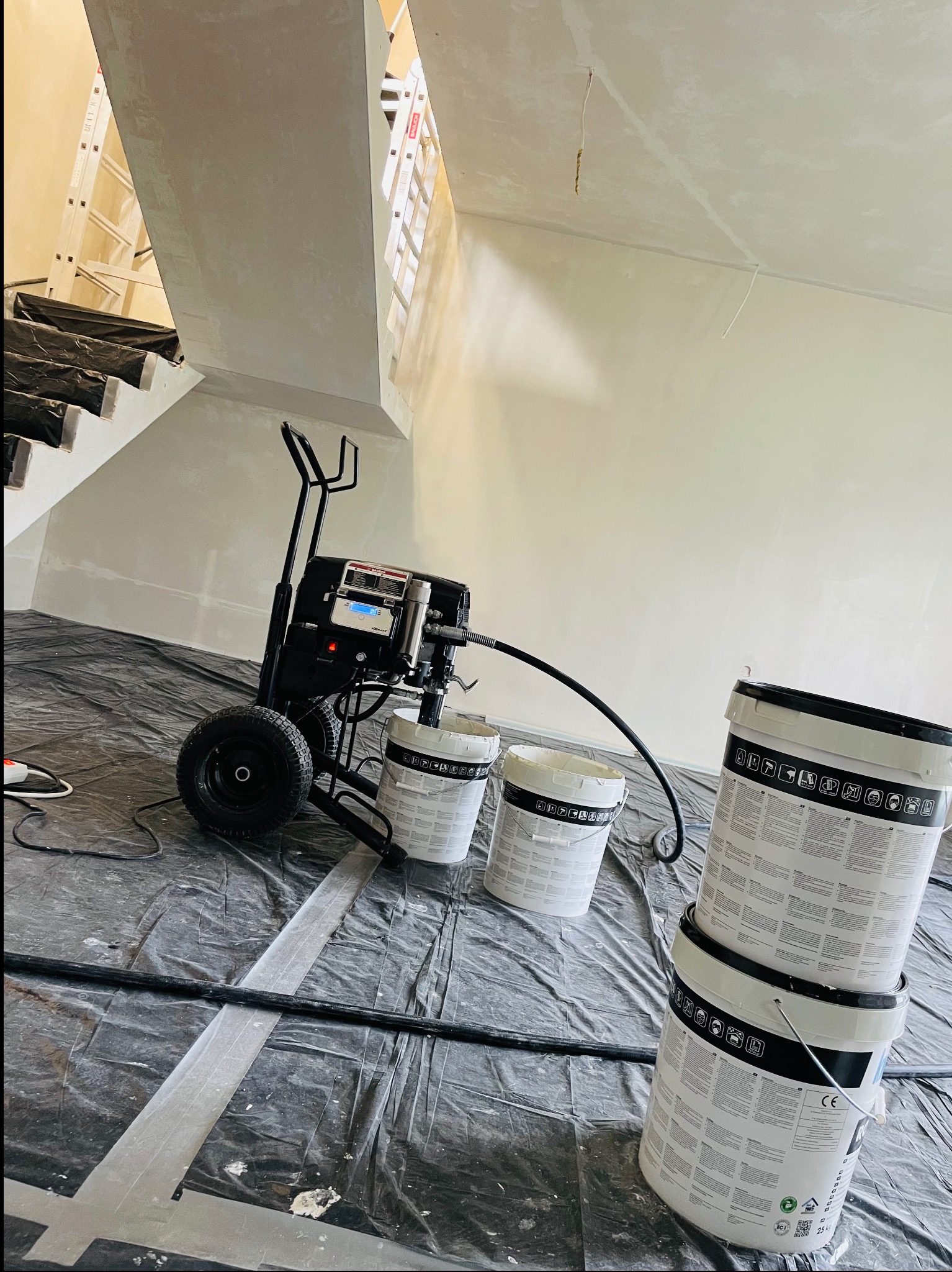 Agregat malarski stoi na folii ochronnej w pomieszczeniu ze schodami, obok ustawione wiadra z farbą, ściany przygotowane do malowania.